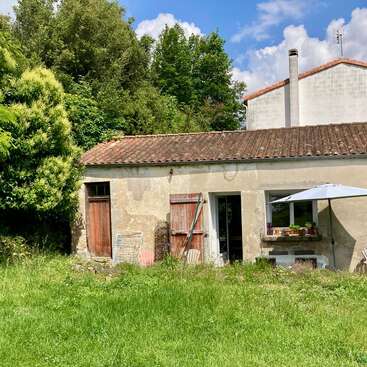 A rustic cottage with red roof tiles stands amid lush greenery, featuring weathered doors, an umbrella, potted plants, and a sunlit, overgrown garden.