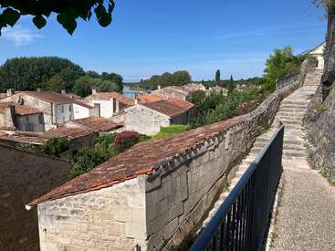 A scenic view of a charming village with stone houses and red-tiled roofs, a stone wall, winding steps, lush greenery, and a river in the distance.