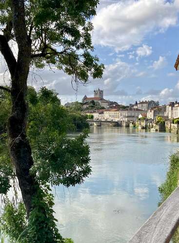 A serene riverside town with a church atop a hill, lined by charming old buildings and lush trees, under a blue sky with fluffy clouds.