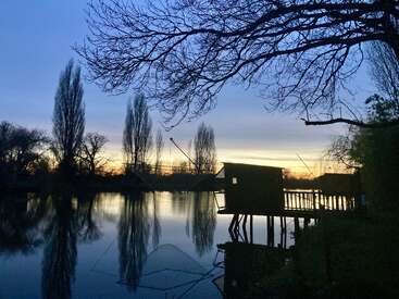 A tranquil riverside scene at dusk, featuring a silhouetted fishing shack on stilts, bare trees, calm reflective water, and a softly glowing sunset sky. Peaceful atmosphere.