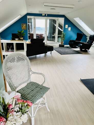 This bright, modern attic room features a white wicker chair with flowers, blue accent wall, cozy black furniture, plants, skylights, and glass doors leading outside.