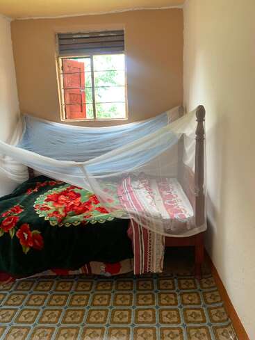 Um quarto pequeno e simples com uma cama de solteiro, cobertor floral, mosquiteiro, piso estampado, paredes creme, janela aberta com persianas vermelhas e luz natural entrando.