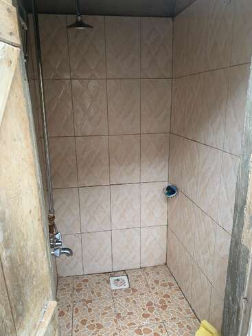 É um chuveiro pequeno, revestido com azulejos, com um chuveiro básico, torneira e ralo. Os azulejos bege cobrem as paredes e os mosaicos cobrem o piso. Design rústico e simples.