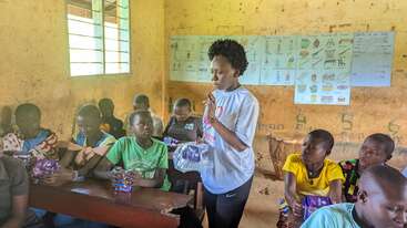 Um professor está em pé na sala de aula distribuindo pacotes roxos para os alunos atentos, sentados nas carteiras. Cartazes educativos cobrem a parede. A luz do sol entra pela janela.