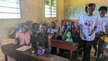 Um grupo de alunos numa sala de aula segurando pacotes, com funcionários ao lado. A sala tem paredes amarelas, pôsteres e a luz do sol entrando pelas janelas. Sorrisos e interação.