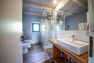 Este moderno cuarto de baño cuenta con una ducha de cristal, tocador de madera, lavabo de recipiente, azulejos blancos, bidé, inodoro, una pequeña ventana, plantas y decoración náutica bajo un techo de vigas.