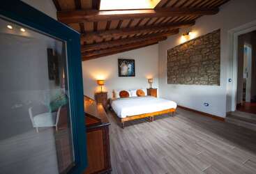 Este acogedor dormitorio cuenta con techo de madera, pared de piedra, cama de matrimonio, dos lámparas, muebles rústicos, suelo moderno y un ambiente luminoso y acogedor.