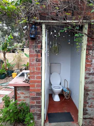 La imagen muestra un aseo exterior con un inodoro y un lavabo blancos, rodeado por un muro de ladrillo y un jardín, con una puerta y una mesita con un farol.