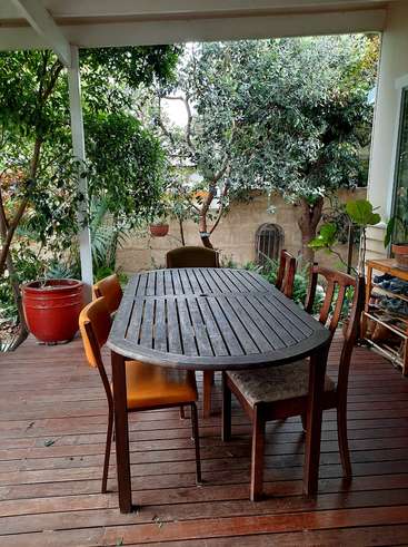 La imagen muestra un sereno patio exterior con una mesa y sillas de madera oscura sobre una terraza de madera, rodeado de exuberante vegetación y un muro de piedra al fondo.