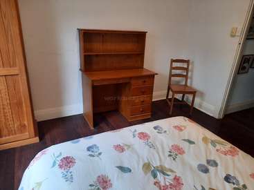 La imagen muestra un dormitorio con cama, escritorio y silla, con edredón de motivos florales y muebles de madera sobre una pared blanca con suelo de madera oscura.