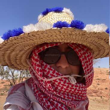 Eine Person, die einen großen Strohhut mit blauen und weißen Verzierungen, eine Sonnenbrille und einen rot gemusterten Schal trägt, der ihr Gesicht bedeckt, steht in einer sonnigen Wüstenlandschaft.