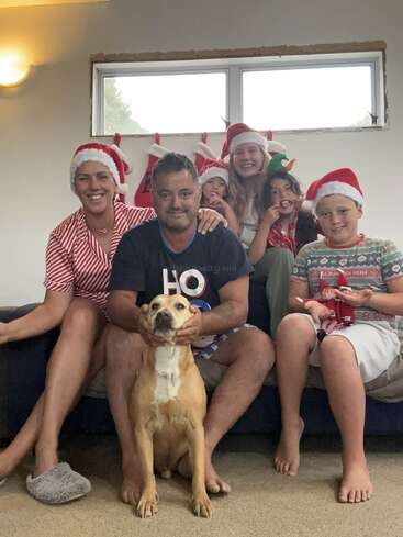 Uma família de seis pessoas, incluindo um cachorro, posa alegremente dentro de casa usando chapéus de Natal. Meias estão penduradas atrás deles. Todos sorriem, criando uma atmosfera festiva e acolhedora de fim de ano.