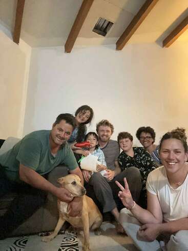 Um grupo de seis pessoas e um cachorro posam juntos em uma sala aconchegante, sorridentes e felizes. Uma pessoa faz o sinal da paz; todos parecem alegres.