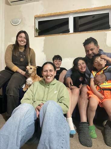 Uma família feliz, incluindo um cachorro, posa junta dentro de casa. O quarto parece estar em reforma. Todos estão sorrindo, criando uma atmosfera calorosa e alegre.
