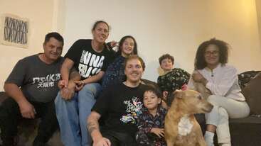 Um grupo feliz de sete pessoas, incluindo adultos e crianças, senta-se junto num sofá dentro de uma sala de estar aconchegante, com um cão castanho à frente.