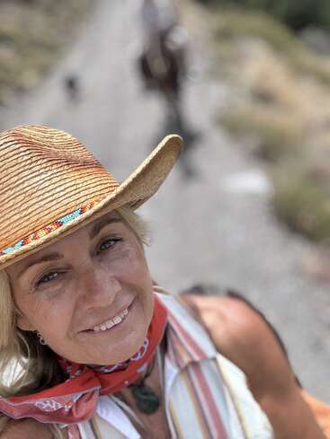 Eine lächelnde Frau mit Strohhut und rotem Halstuch macht ein Selfie im Freien auf einem Wanderweg, mit unscharfem Reiter und Pferd im Hintergrund. Ein heller, sonniger Tag.