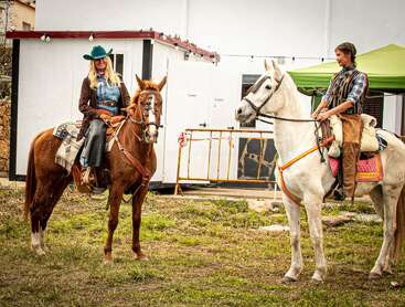 Zwei als Cowboys gekleidete Personen sitzen auf Pferden und stehen sich gegenüber. Sie befinden sich im Freien, in der Nähe eines weißen Gebäudes und einer grünen Überdachung auf Gras.