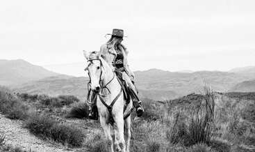 A woman wearing a hat rides a white horse through a rugged landscape. The scene is in black and white, evoking a sense of adventure.