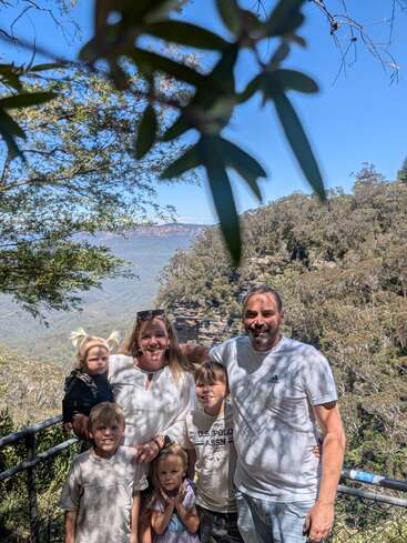 Eine sechsköpfige Familie lächelt an einem sonnigen Tag im Freien, umgeben von Bäumen und Bergen, an einem malerischen Aussichtspunkt mit blauem Himmel.