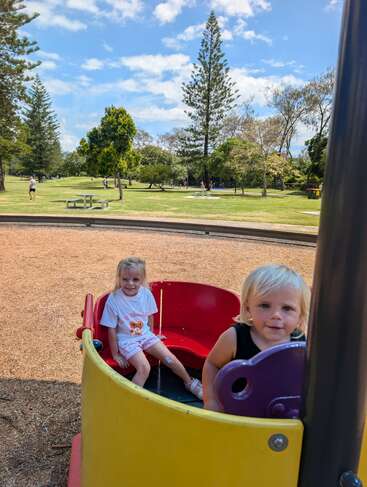Zwei Kinder spielen fröhlich auf einem bunten Spielplatzkarussell. Hinter ihnen liegt ein malerischer Park mit Bäumen, Wiesen, Picknickbänken und Menschen, die die Natur genießen.