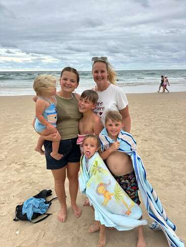 Eine Frau und vier Kinder stehen lächelnd an einem Sandstrand. Das Meer und der bewölkte Himmel liegen hinter ihnen. Zwei Kinder sind in Handtücher gewickelt und haben Spaß.