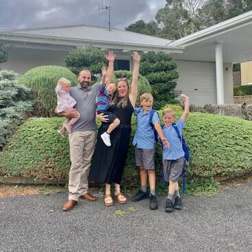 Una alegre familia de cinco miembros posa delante de su casa, con los padres sosteniendo a dos niños y tres niños emocionados, con los brazos levantados, listos para un día de colegio.