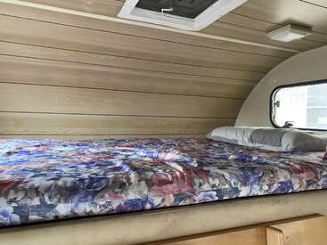 This cozy camper interior features a floral-patterned bed, wood-paneled walls and ceiling, a small window, pillows, and a skylight, creating a warm, inviting space.