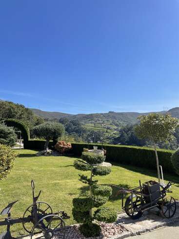The image depicts a serene garden with a manicured lawn, trimmed trees, and a vintage cart, set against a backdrop of rolling hills and a clear blue sky.