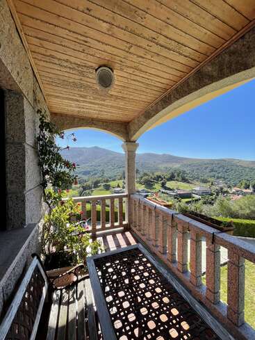 The image depicts a serene balcony with a wooden ceiling and railing, overlooking a picturesque landscape of rolling hills and mountains under a clear blue sky.