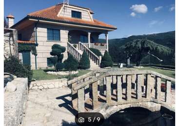 The image depicts a two-story house with a stone bridge over a pond, surrounded by trees and a mountainous landscape under a clear blue sky.