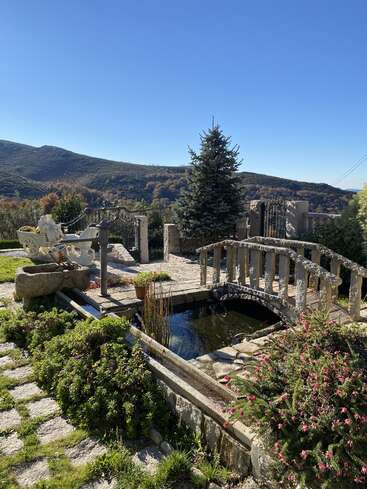 The image depicts a serene garden with a stone bridge, pond, and fountain, set against a backdrop of rolling hills and a clear blue sky.