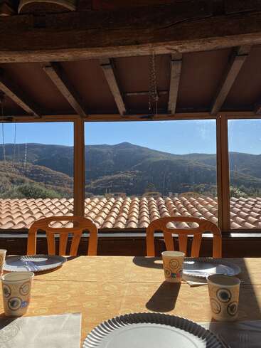 The image depicts a rustic dining room with a table set for a meal, featuring a scenic view of rolling hills and mountains through large windows.