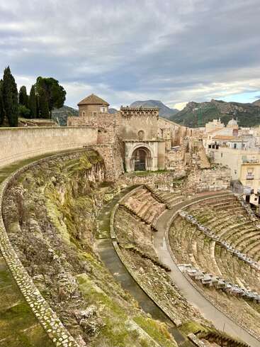 This image shows an ancient Roman amphitheater with stone seating, partially covered in moss, surrounded by historical buildings, cypress trees, and distant mountains under cloudy skies.