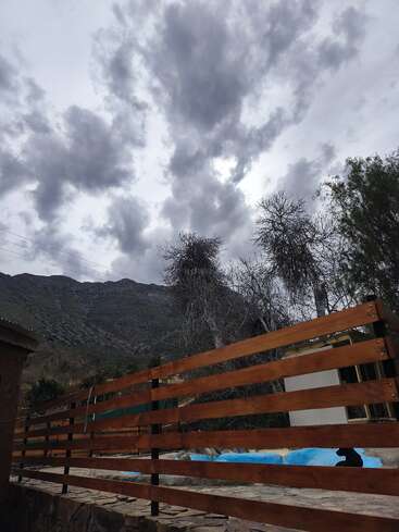 Des nuages sombres remplissent le ciel au-dessus des montagnes. Une clôture en bois et un chemin en pierre se trouvent au premier plan, avec des arbres dénudés et un peu de verdure à l'arrière-plan.