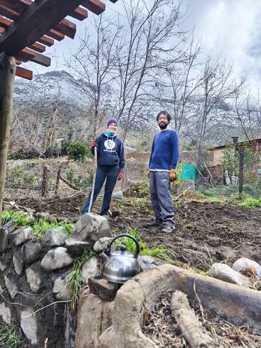 Two people stand on soil in a garden, surrounded by trees and mountains. A kettle sits on a stone wall. The sky is cloudy and gray.