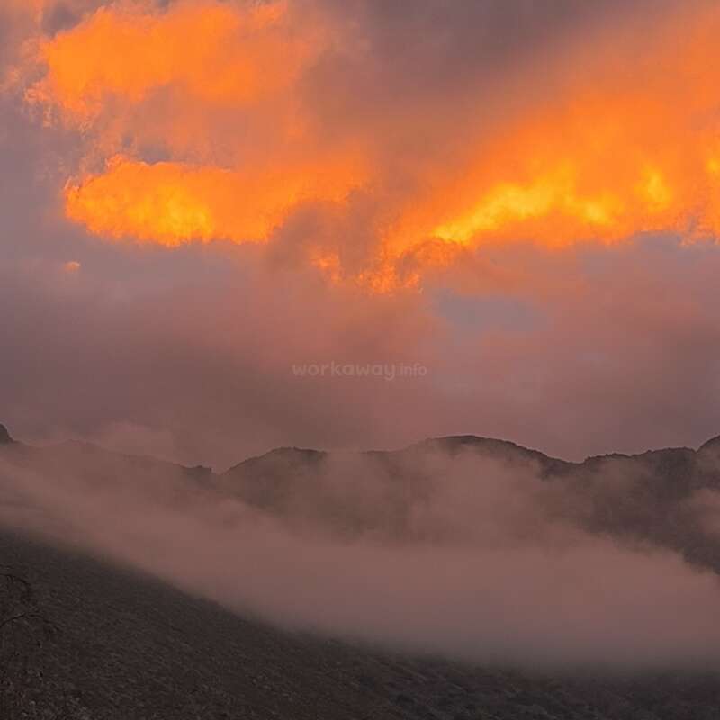 Un ciel orange et jaune dramatique s'illumine au coucher du soleil au-dessus de montagnes silhouettées, tandis que la brume et le brouillard roulent doucement sur le paysage sombre et ombragé en contrebas. Atmosphère paisible.
