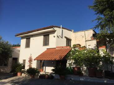 This image shows a Mediterranean-style house with white walls, a red-tiled roof, black window grilles, potted plants, lush greenery, and clear blue sky above.