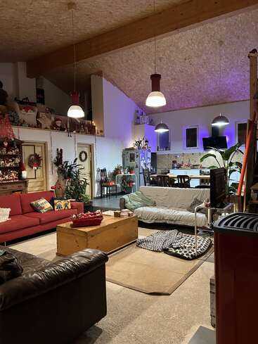 A cozy, eclectic living space decorated for Christmas. Features comfortable sofas, wooden beams, hanging lights, a dining area, plants, and a festive, welcoming atmosphere.