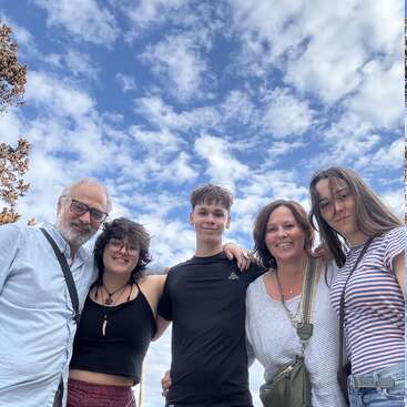 A happy family of five stands closely together, smiling under a beautiful blue sky filled with scattered clouds. They look relaxed, enjoying a day outdoors together.