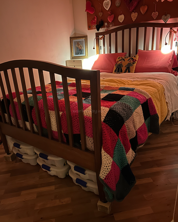 Cozy bedroom with a wooden bed, colorful crocheted blanket, yellow comforter, pink pillows, heart decorations, bedside painting, and storage containers under the bed. Warm lighting.