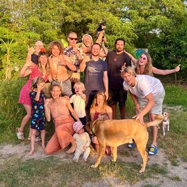 A joyful group of adults, children, and dogs pose together outdoors, surrounded by lush greenery, smiling and making playful gestures in the warm, golden sunlight.