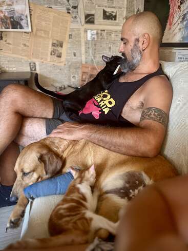 Un hombre sentado en un sofá con un gato negro en su regazo, acariciando a un perro dormido con una escayola y un gatito a su lado. Una escena acogedora.