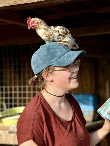 Una persona sonriente con gafas y gorra azul está fuera, sosteniendo una lata, con una gallina moteada cómodamente posada en su sombrero, disfrutando del momento.