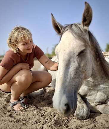 Una joven de pelo corto y rubio acaricia suavemente a un relajado caballo gris tumbado en el suelo arenoso, ambos disfrutan juntos de un momento de paz y sol al aire libre.
