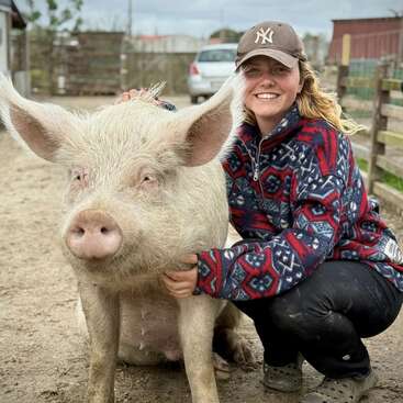 A smiling woman wearing a baseball cap and patterned jacket kneels beside a large, friendly pig outdoors on a farm, showcasing a joyful human-animal bond.