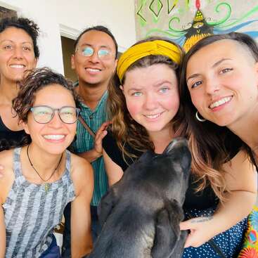 The image shows five people and a dog posing for a selfie, with the dog in the foreground and the group standing together in front of a wall with colorful artwork.
