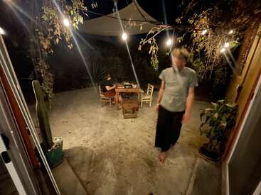 Deux personnes sont installées sur une terrasse confortable, en soirée. Des guirlandes lumineuses sont suspendues au-dessus de leurs têtes et éclairent une table garnie de plats. Des plantes entourent l'espace, créant une ambiance chaleureuse.