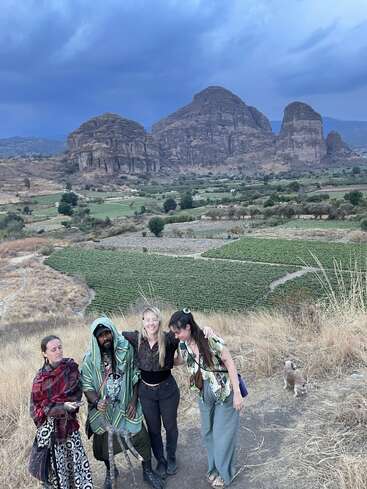 Quatre personnes et un petit chien se tiennent sur le flanc d'une colline, avec en toile de fond des montagnes rocheuses impressionnantes et des terres agricoles verdoyantes, le tout sous un ciel nuageux spectaculaire.