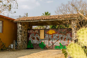Un portail rustique, avec ses piliers en pierre et son toit en tuiles, est orné de graffitis colorés, de cactus et d'arbres. La lumière du soleil projette des ombres, créant une atmosphère chaleureuse dans ce décor champêtre.