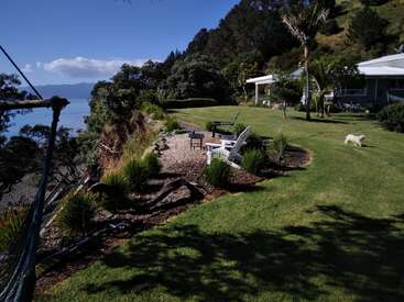 Un hermoso jardín costero cuenta con sillas Adirondack alrededor de una hoguera. El césped verde, las vistas al mar, la hamaca, los árboles y un pequeño perro blanco realzan la tranquilidad del lugar.